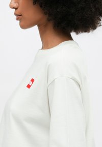 Heledad kergelt kreemjas puuvillas, millel on mugav lõige. Ees on väike punane logo plaat rinna kohal ja laiad, veidi rullitud varrukad.