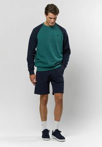 Tumši zaļš džemperis ar tumši ziliem raglan piedurknēm, savienots ar tumši ziliem šortiem. Modelis valkā baltas ribotas zeķes un tumši zilus sporta apavus.