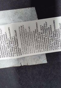 White fabric care label showing various languages, detailing washing instructions; text printed in black, accompanied by a textured, frayed edge.
