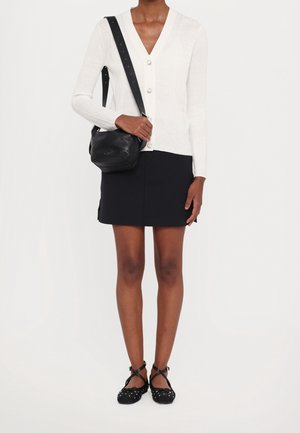 Vrouw met een wit vest met knopen, een zwarte minirok, een zwarte crossbodytas en zwarte enkellaarsjes met studs, staand tegen een witte achtergrond.