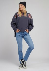 Tummanharmaa huppari, jossa on rohkea "RIDER"-teksti, yhdistettynä vaaleisiin farkkuihin ja mustiin korkeavartisiin tennareihin. Beanie viimeistelee rennon ilmeen.