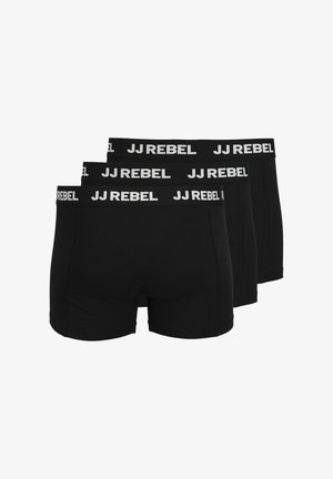 Jeune homme portant un boxer noir JJ Rebel, debout devant un fond clair uni, ajustant la ceinture avec une main.