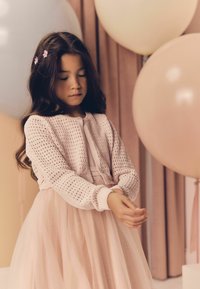 Cardigan lavorato a maglia rosa chiaro con chiusura frontale, materiale texturizzato, abbinato a un vestito di tulle rosa a strati; ragazza con capelli lunghi e scuri e fermagli floreali nei capelli.