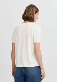 T-shirt blanc à manches courtes avec des manches ballon, en tissu texturé, et une finition décorative autour de l'encolure et des manches.