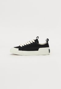 Sorte lerret sneakers med kremfargede lisser, gummitåhett og teksturert hvit såle. Inkluderer et merke på baksiden med en logo.