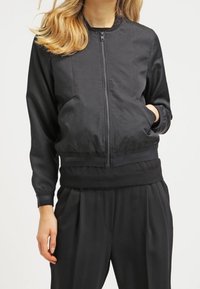 Czarna kurtka bombowca z suwakowym przodem, ściągaczami przy mankietach i w pasie. Wykonana z gładkiego materiału, posiada boczne kieszenie. Połączona z pasującymi spodniami.