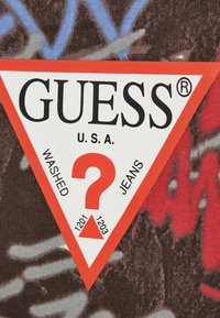 Etichetta triangolare rossa e bianca con testo nero, che presenta le parole "GUESS," "WASHED," "JEANS," e un punto interrogativo. Sfondo in tessuto a fantasia.