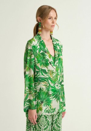 Femme aux cheveux clairs portant un blazer à motif feuillu vert et un pantalon à motif vert, accessoirisée avec de longues boucles d'oreilles à pampilles multicolores.