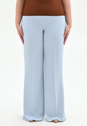 Femme portant un pantalon large bleu clair et un top côtelé marron, debout avec les mains le long du corps sur fond blanc.