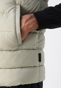Light grey puffer vest with vertical stitching and a side pocket. Features a small green tag reading “EST. 1993” near the hem.