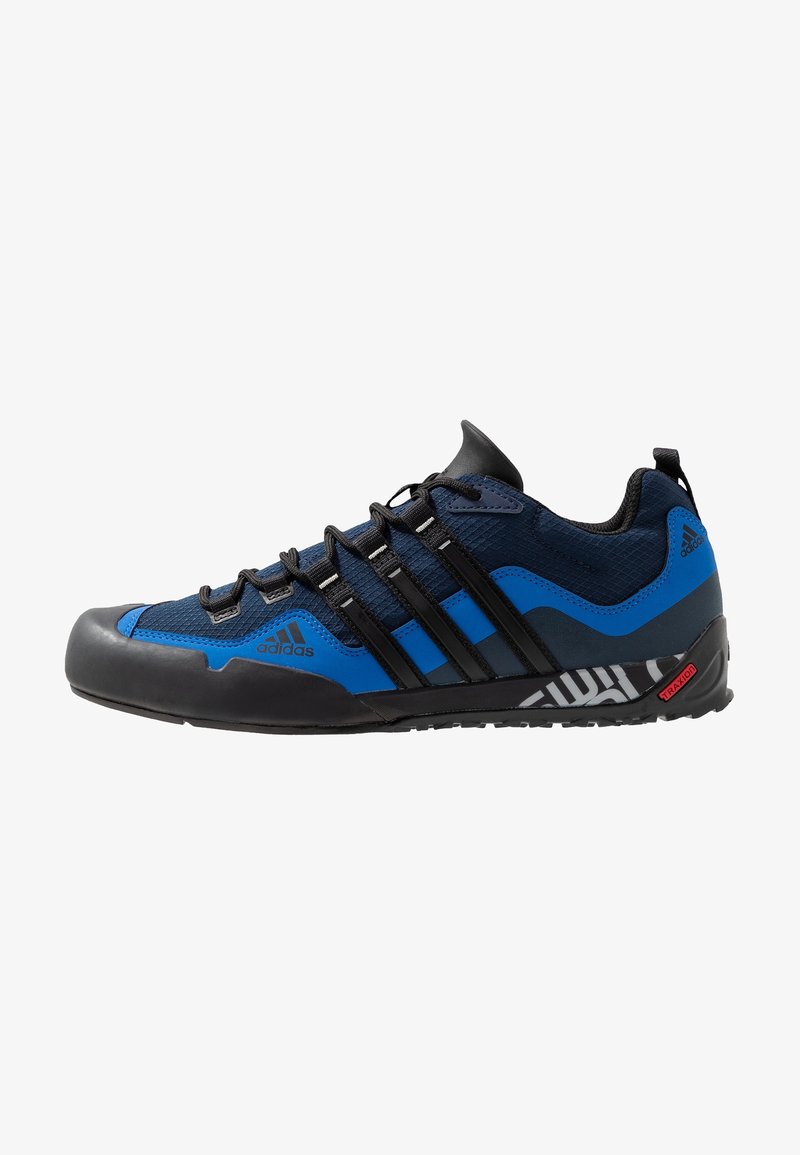 Chaussure de sport Adidas basse bleue et noire avec lacets noirs, trois bandes noires sur les côtés, et semelle en caoutchouc texturée avec étiquette Traxion.