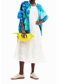 Osoba oblečená v bílých vrstvených šatech, modrozelené vzorované bundě, béžových teniskách, držící jasně žlutou kabelku s logem "Desigual".