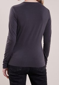 Maglia aderente a maniche lunghe in tessuto grigio scuro con una texture liscia e scollo rotondo, abbinata a jeans scuri. Nessun motivo o accessorio visibile.