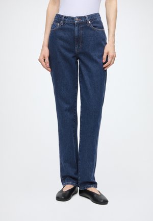 Person iført mørkeblå straight-leg jeans, hvid top og sorte flade sko, der står mod en ensfarvet lys baggrund.