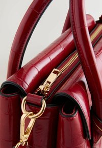 Rote, glanzende Handtasche mit Krokodilstruktur, versehen mit goldenen Beschlägen, einem Reißverschluss und zwei Griffen. Akzente setzen schwarze Kanten.
