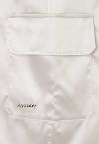 Tissu blanc satiné avec une poche, arborant un logo marron foncé. Design simple avec une texture lisse, mettant en valeur les détails de couture.