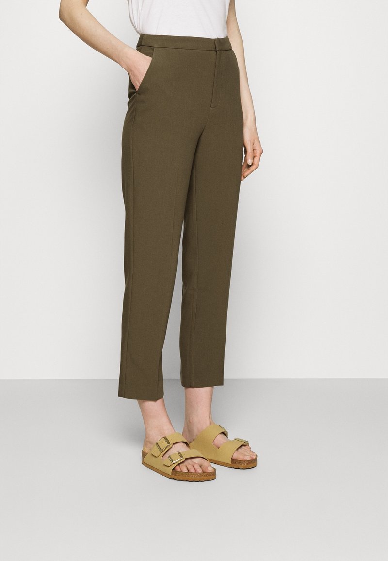 Personne portant un pantalon droit court vert olive, un haut blanc et des sandales beiges à double bride avec boucles, debout devant un fond blanc.