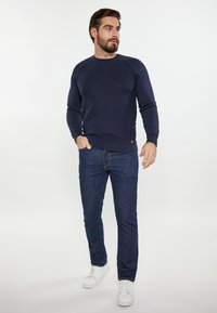 Marineblauwe geribbelde trui met lange mouwen en ronde hals, gecombineerd met donkere spijkerbroeken en witte sneakers. Bevat een klein logo.