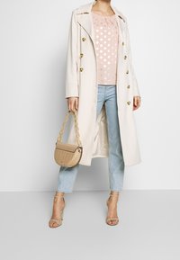 Ljusbeige trenchcoat med guldknappar över en rosa prickig topp. Avslappnade blå jeans, en beige handväska och klackar med remmar kompletterar outfiten.