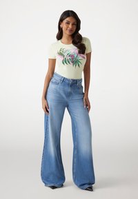 T-shirt jaune clair à manches courtes avec un design graphique floral ; associé à un jean en denim bleu à jambe large avec une finition délavée et des poches avant.