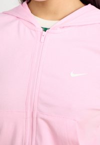 Närbild av en person iklädd en rosa zip-up hoodie med en vit Nike-logotyp på bröstet och en grön t-shirt under.