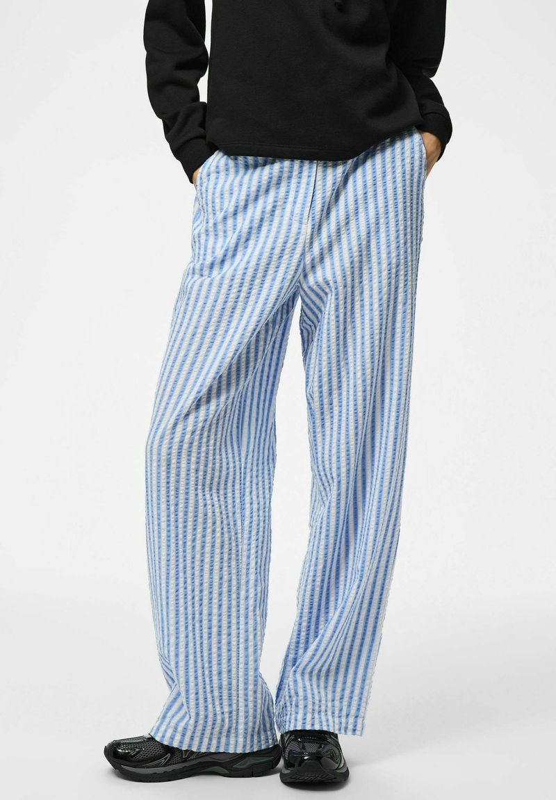 Personne portant un pantalon ample à rayures verticales bleues et blanches, un sweat-shirt noir et des baskets noires, debout les mains dans les poches.