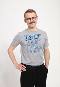 Camiseta de algodón gris con una ilustración de caricatura y el texto "¡EL DESTINO TE ESPERA!" en azul, combinada con pantalones negros y un cinturón.