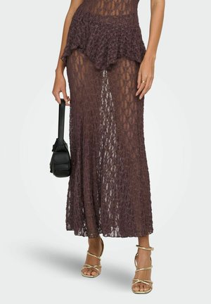 Kobieta nosząca przezroczystą ciemnobrązową koronkową sukienkę, trzymająca czarną torebkę, w połączeniu ze złotymi sandałami na wysokim obcasie z paskami, na jednolitym tle.