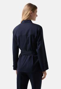 Navy fabric jacket with a belted waist, long sleeves, and a back yoke detail. Smooth texture and buttoned cuffs add notable accents.