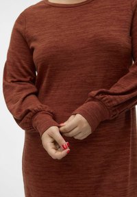 Abito a maniche lunghe color ruggine con tessuto testurizzato, caratterizzato da polsini arricciati e scollatura tonda. Messa a fuoco sulle mani che sistemano le maniche.
