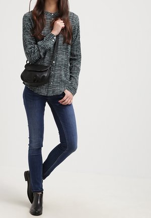 Nainen, jolla on kuvioitu pitkähihainen paita, tummansiniset kapealahkeiset farkut, mustat nilkkurit ja kädessään mustan olkalaukun hihna.