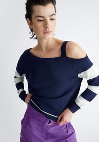 Maglione a maglia navy con design off-shoulder, dettagli a righe bianche sulle maniche e collo a coste. Indossato con pantaloni cargo viola.
