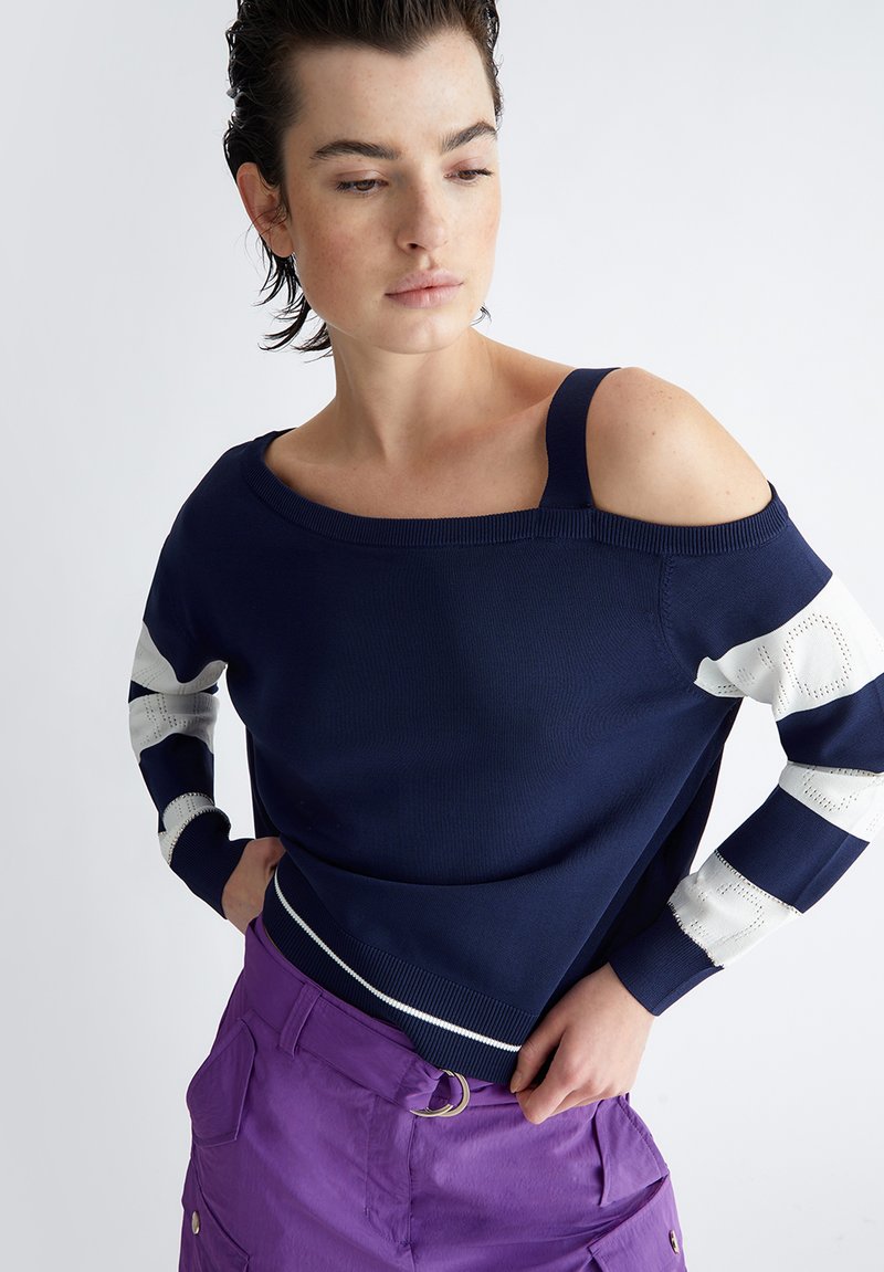 Maglione a maglia navy con design off-shoulder, dettagli a righe bianche sulle maniche e collo a coste. Indossato con pantaloni cargo viola.