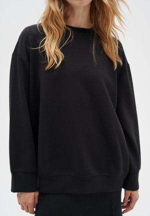 Woman with blonde hair wearing a loose black long-sleeve sweatshirt and a dark skirt, standing against a plain background.