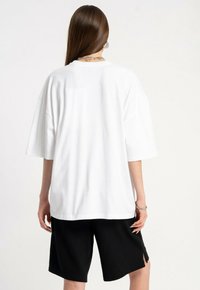 Osoba z długimi brązowymi włosami, ubrana w białą, dużą koszulkę oraz czarne spodenki do kolan, stojąca tyłem do aparatu.