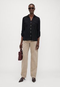 Chemise noire à manches longues avec deux poches poitrine, pantalon beige taille haute, sac à main bordeaux et chaussures brunes à bout pointu, fond neutre.