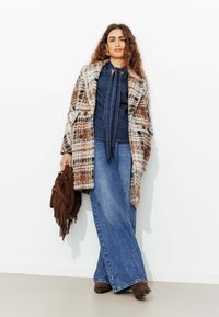 Manteau en laine à carreaux marron et blanc, haut en denim, jeans évasés et sac en suédine marron à franges. Des bottes à talons marron complètent la tenue.