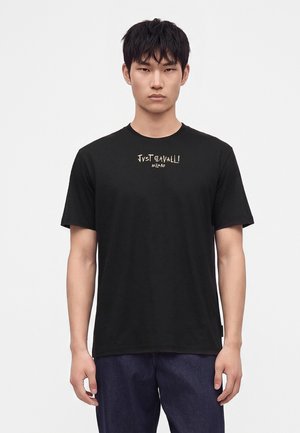 Ung mand iført en sort Just Cavalli Milano t-shirt og mørke bukser, står foran en ensfarvet hvid baggrund.