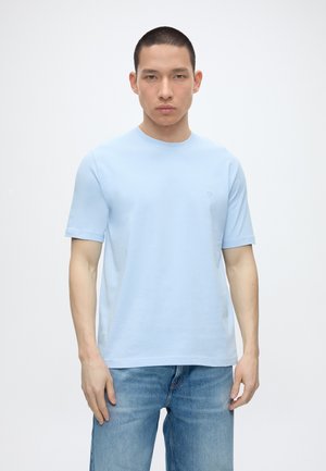 Muž oblečený v svetlomodrom tričku s krátkym rukávom a okrúhlym výstrihom a modrých džínsoch, stojaci pred bielym pozadím s neutrálnym výrazom.