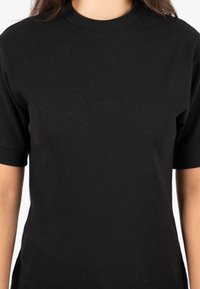 Donna che indossa una maglietta nera a costine a maniche corte, semplice, con un piccolo logo ricamato e discreto al centro del petto.