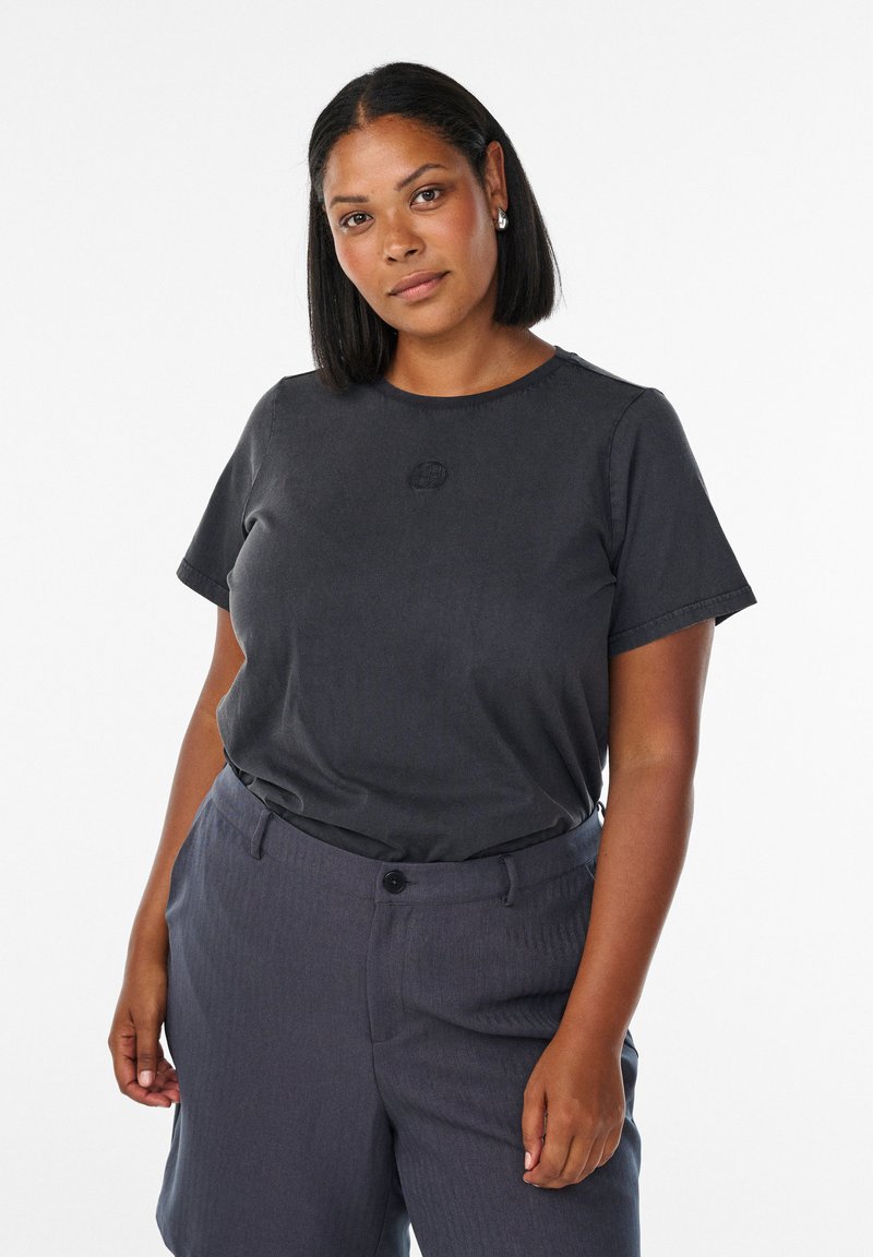 Kvinde med skulderlangt sort hår iført en mørkegrå T-shirt og marineblå bukser, der står mod en ensfarvet hvid baggrund.