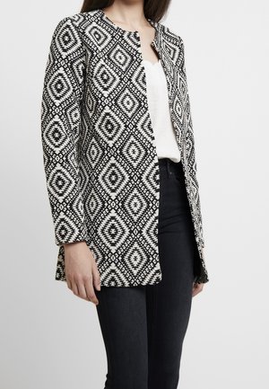 Woman wearing a black and white geometric patterned long-sleeve jacket over a white top and black pants, standing against a plain background.