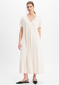 Maxikleid aus leicht beigem Stoff, V-Ausschnitt, kurzen Ärmeln und plissiertem Frontdetail. Kombiniert mit schwarzen spitzen Ballerinas. Glatte Textur.