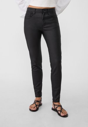 Femme portant un pantalon skinny noir et des sandales plates noires à brides, debout devant un fond clair uni.