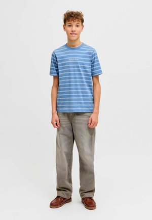 Pusaudzis zēns stāv, raugoties taisni priekšā, valkājot zilu svītrainu T-kreklu, vaļīgas pelēkas džinsa bikses un brūnas čības, fonā ir vienkāršs balts fons.