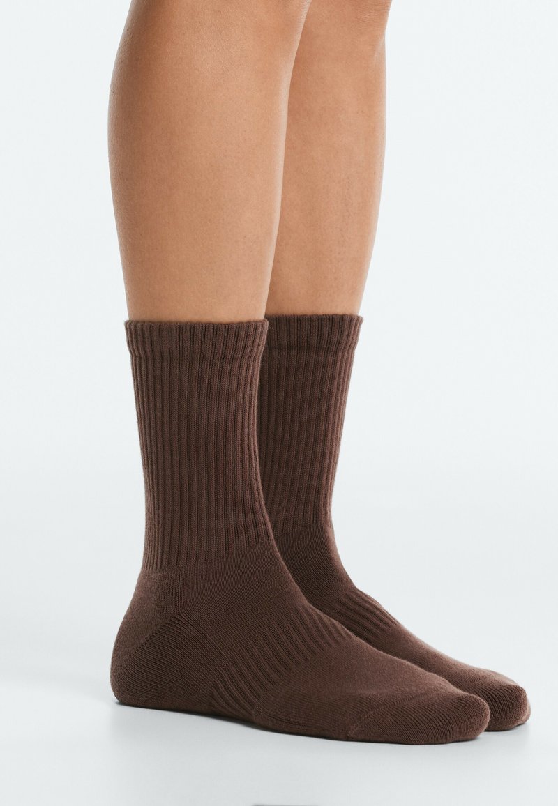 Jambes portant des chaussettes montantes côtelées marron sur un fond clair uni, montrant la texture et l'ajustement des chaussettes.