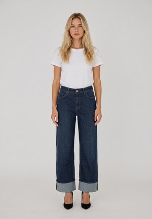 Mujer de pie con camisa blanca de manga corta, vaqueros anchos y oscuros con puños, y zapatos de tacón negros sobre un fondo claro y liso.