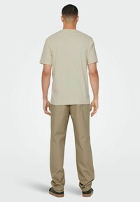 Beige T-Shirt mit kurzen Ärmeln, kombiniert mit hellbraunen Hosen. Der Stoff wirkt weich und hat eine lässige Passform, getragen mit schwarzen Slippern.