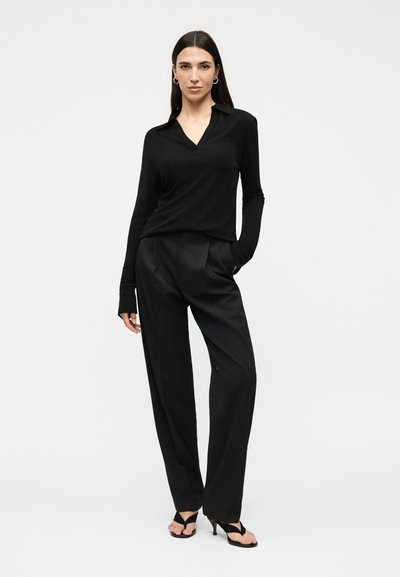 Γυναίκα με μακριά σκούρα μαλλιά, φοράει μαύρο μακρυμάνικο τοπ με λαιμόκοψη V, μαύρο παντελόνι με φαρδιά μπατζάκια και μαύρα σανδάλια με τακούνι, στέκεται με το ένα χέρι στην τσέπη.