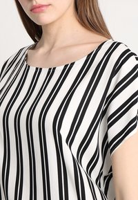 Stribet top i sort og hvid med løstsiddende snit, rund halsudskæring og korte ærmer. Stoffet ser glat ud med en subtil glans.